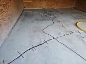 Cracked concrete floor in a wooden-walled room with visible repair wires and cables. The image conveys a sense of construction and repair work.