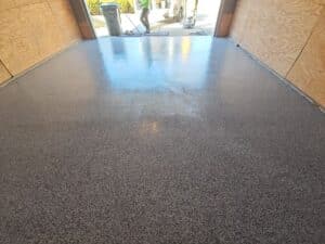 A freshly coated, glossy gray epoxy garage floor stretches to an open door. Wooden walls line the sides, and a worker is seen outside in sunlight.