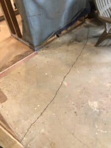 A wide crack running across the bare concrete floor of a living room under renovation, with exposed wall studs and insulation in the background.
