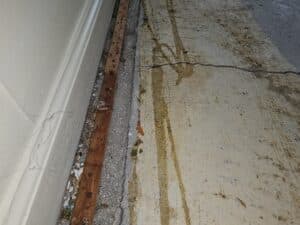 A close-up of a floor corner showing cracked concrete, dirt, and debris near a wooden trim against a white wall.