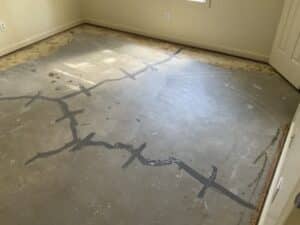 A partially finished concrete floor with visible stains and gray sealing lines from crack stitch in a light-filled room, near a closed door.