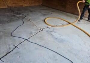 Cracked concrete floor in an empty room; cables and a large orange hose are spread across, held by a person in work boots.