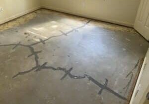 A partially finished concrete floor with visible stains and gray sealing lines from crack stitch in a light-filled room, near a closed door.