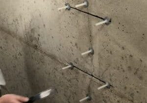 A man in a white coat operates a blue and black tool for epoxy injection for crack stitching, reinforcing basement structures against water entry